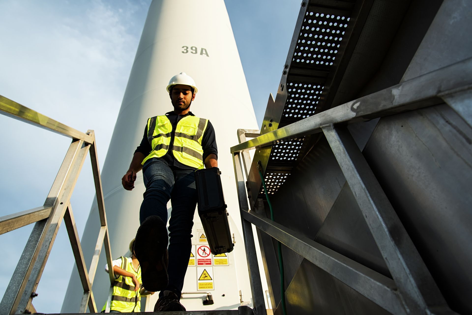 Wind turbine technician descending stairs, carrying a toolbox; wearing safety vest and hard hat. Wind turbine technician descending stairs, carrying a toolbox; wearing safety vest and hard hat.
