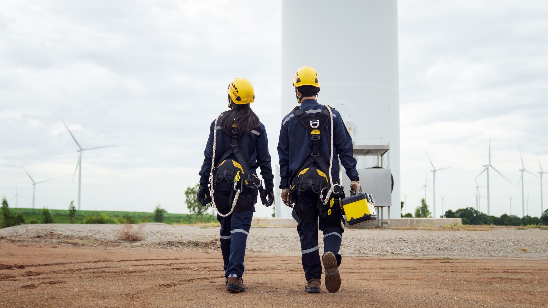 Two people in hard hats and harnesses walk towards a wind turbine, other turbines in the background.