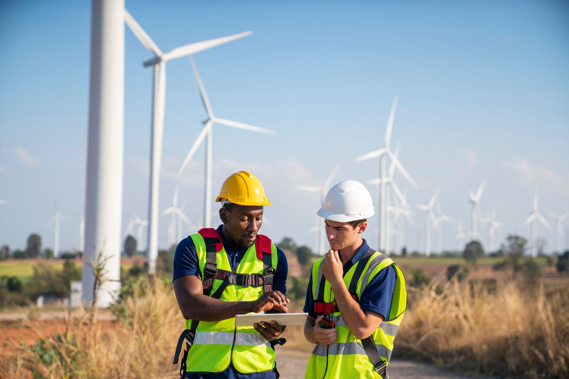 Two engineers reviewing data on a tablet, wind turbines in the background. Two engineers reviewing data on a tablet, wind turbines in the background.