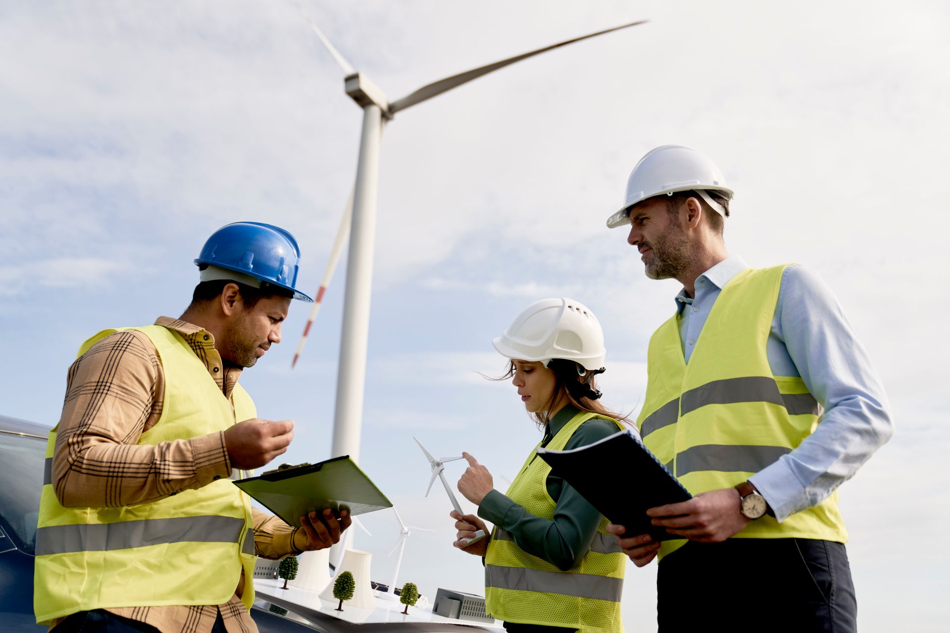 Engineers in hard hats and vests examining models and a wind turbine.