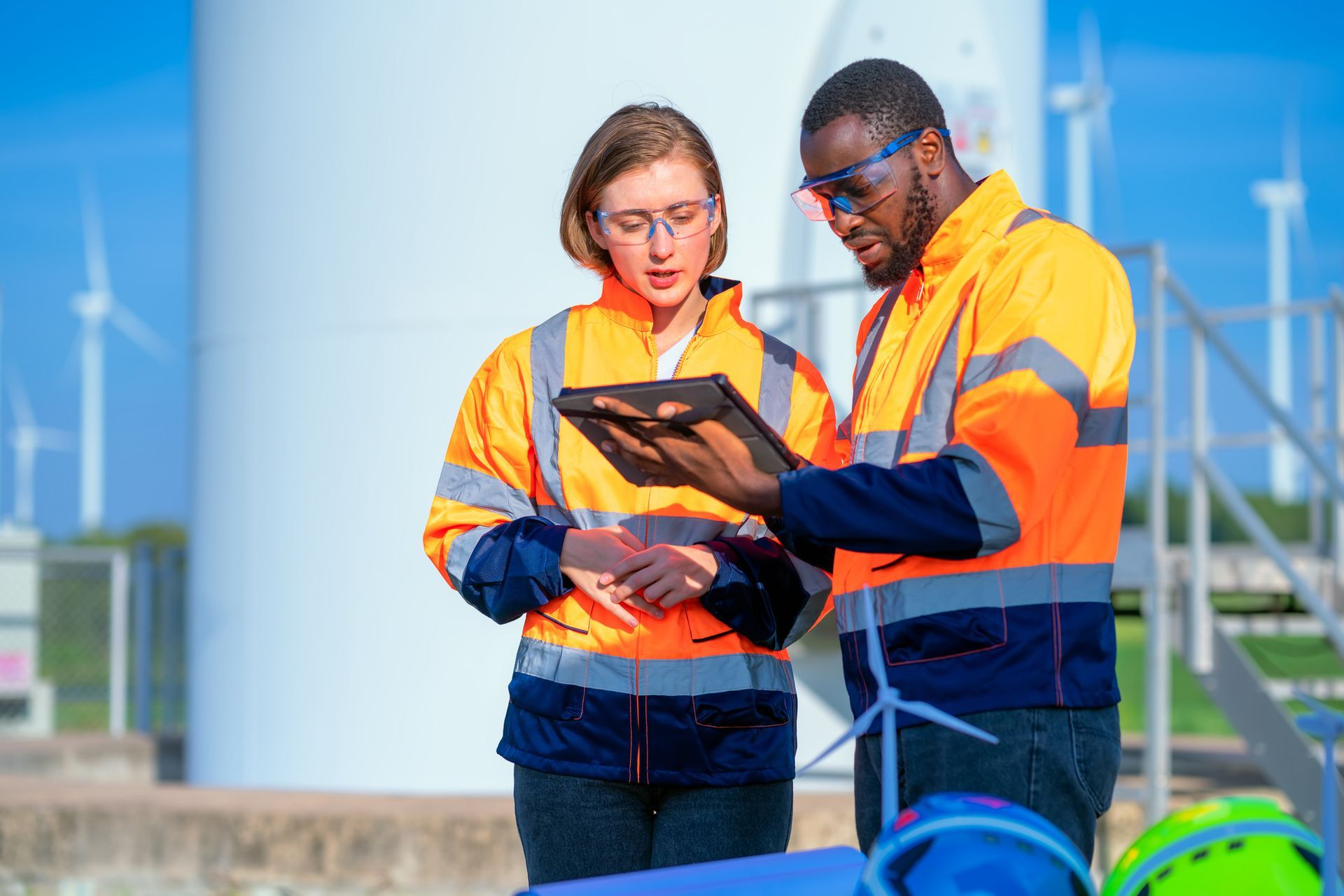 Two engineers in safety vests examining a tablet near wind turbines. Two engineers in safety vests examining a tablet near wind turbines.