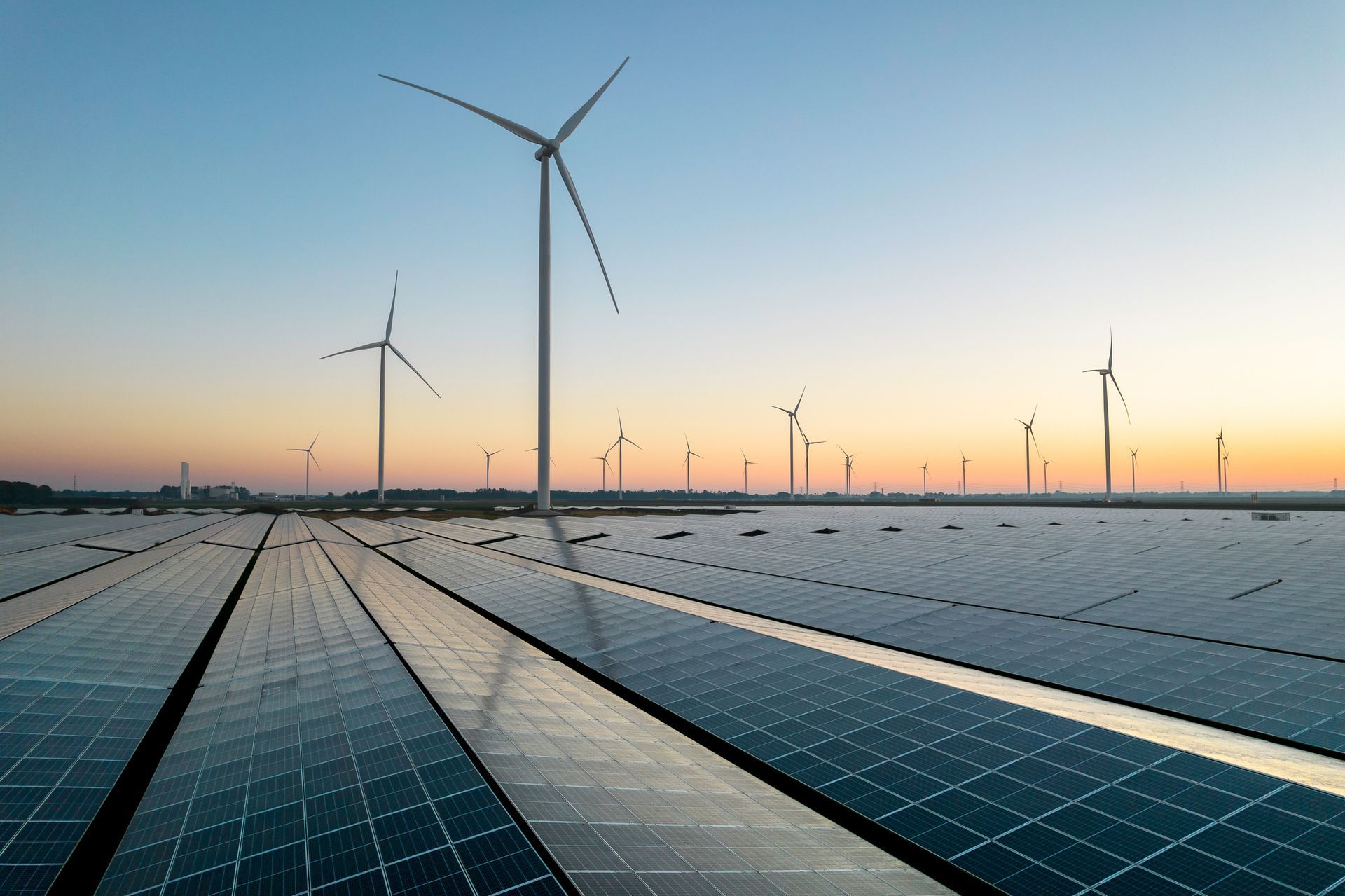 Solar panels with wind turbines in the distance, set against a sunset sky.