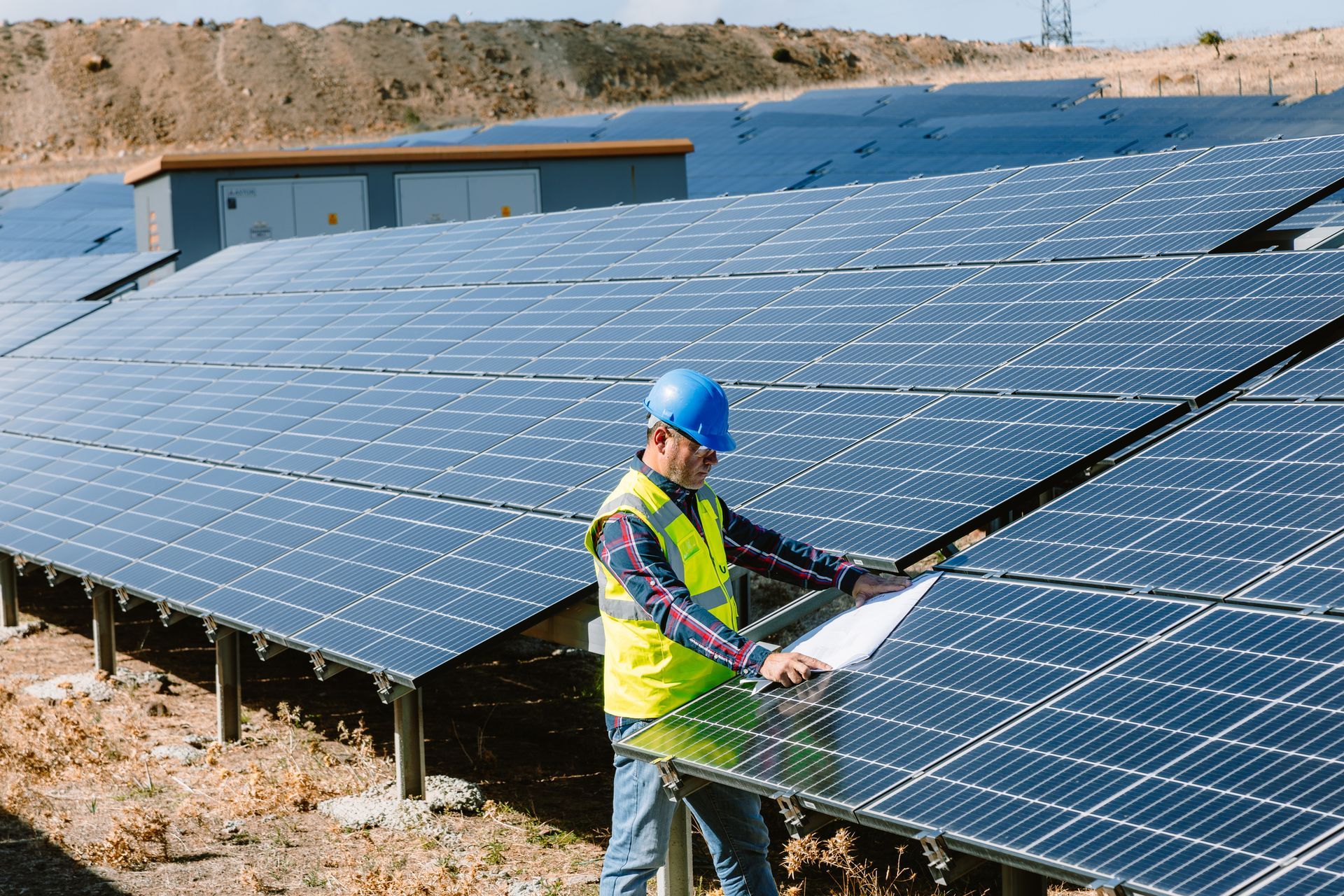 Man in hard hat and safety vest inspecting solar panels outdoors.