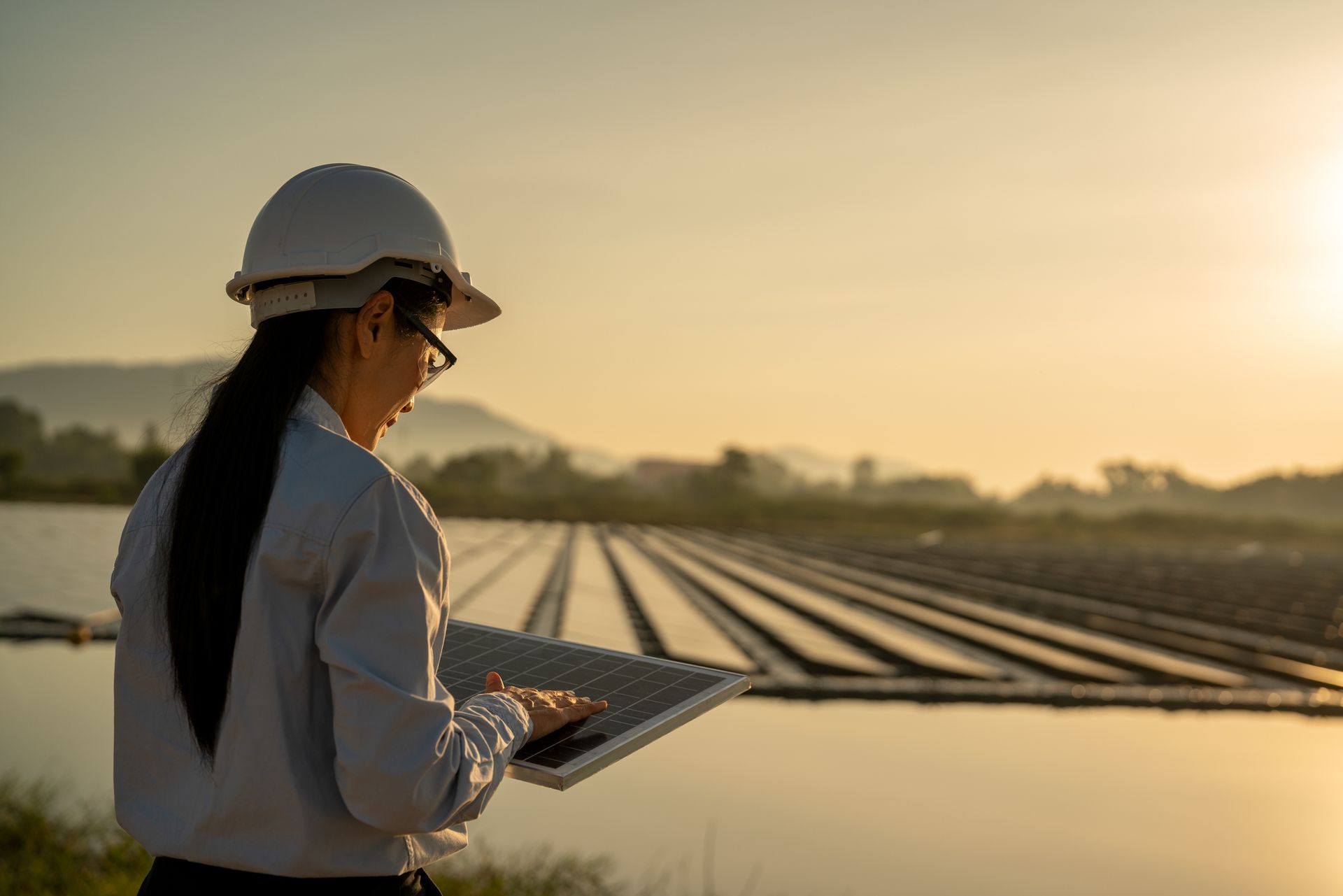 Woman with hard hat and glasses uses a tablet in front of a solar panel field at sunset.