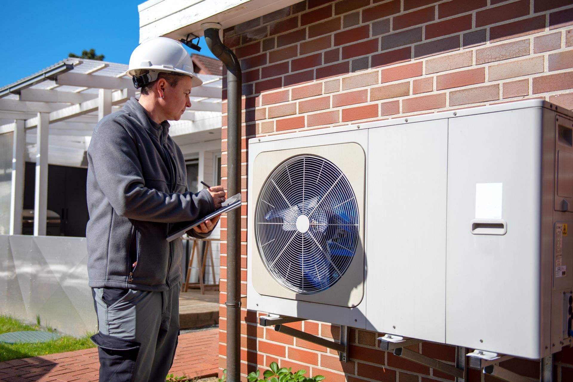 HVAC technician in hard hat inspecting a white air conditioning unit mounted on a brick wall, taking notes.