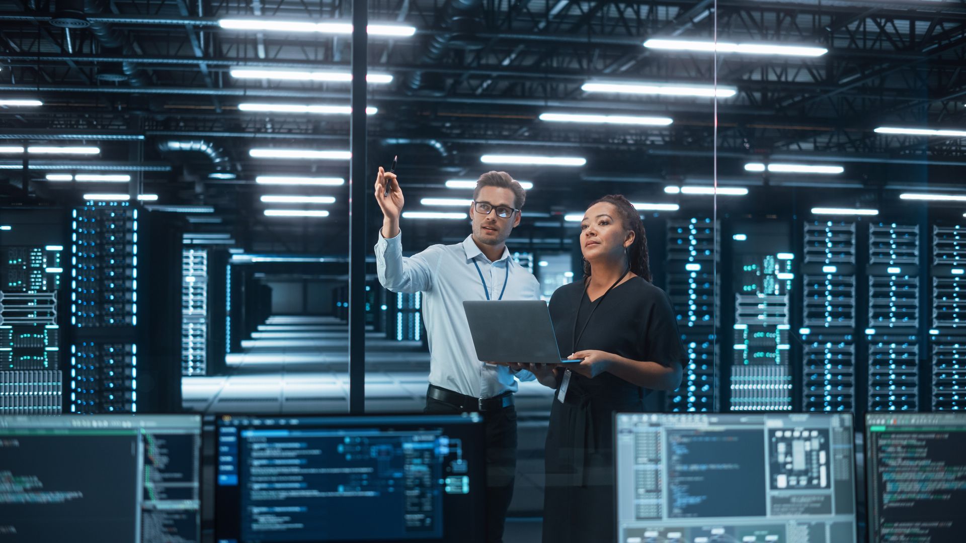 Two people in a data center, man pointing, woman holding laptop, servers in the background, screens in the foreground.