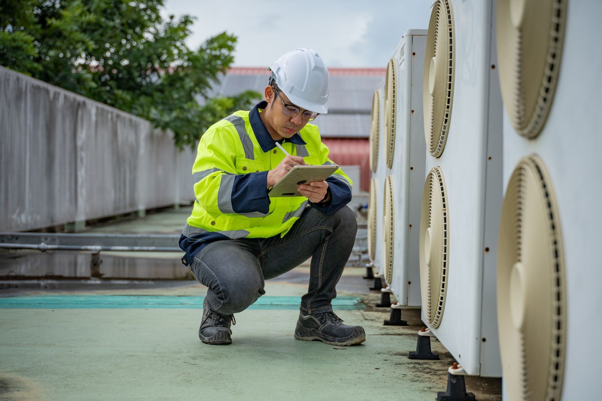 HVAC technician in hard hat and safety vest inspecting rooftop air conditioning units, taking notes on a tablet.