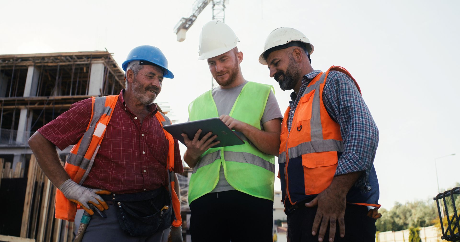Construction workers looking at a tablet on a building site, wearing hard hats and safety vests.
