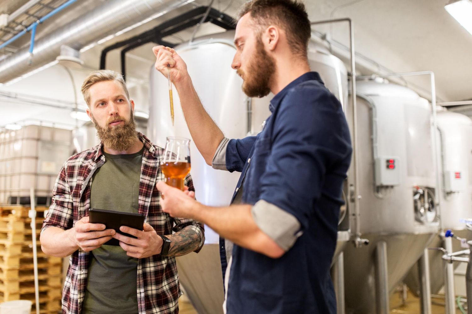 Two men are standing in a brewery looking at a glass of beer.
