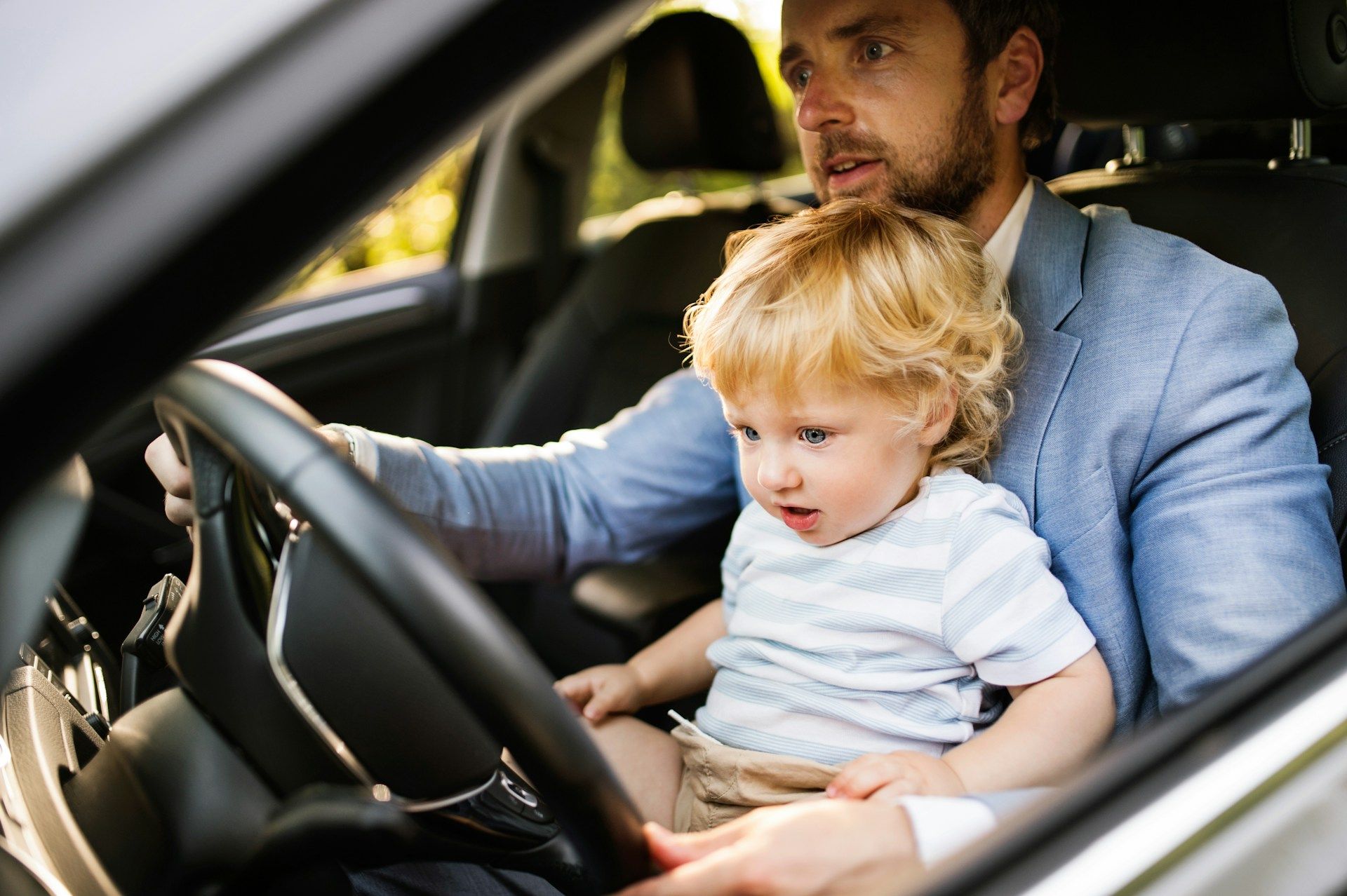 A man is driving a car with a baby in his lap.