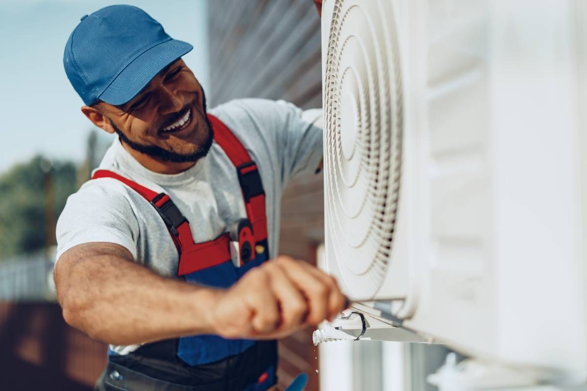 Smiling HVAC professional, wearing a blue hat, repairing an outdoor air conditioner unit near Lexington, Kentucky