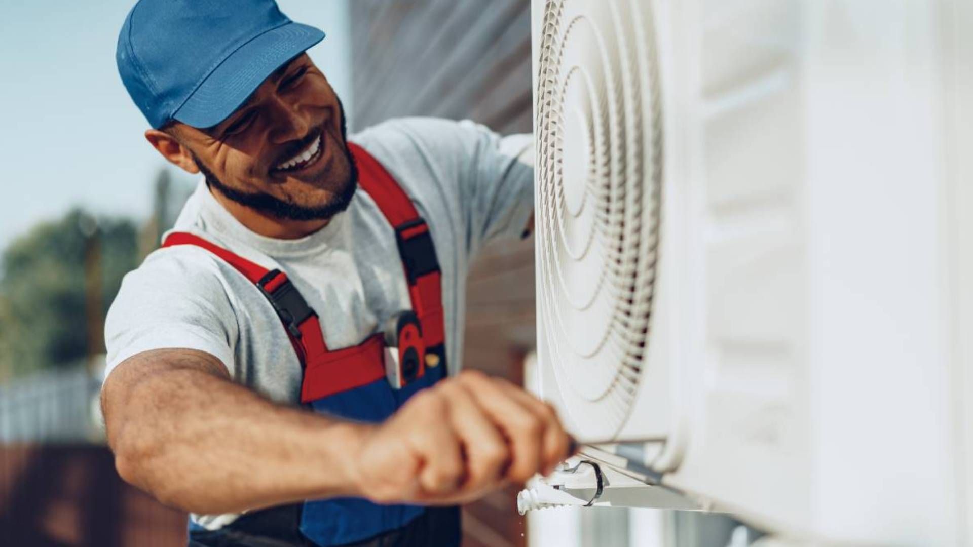 Smiling HVAC professional, wearing a blue hat, repairing an outdoor air conditioner unit near Lexing