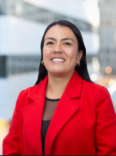 Una mujer de cabello oscuro sonríe, vistiendo un blazer rojo sobre una blusa negra, sobre un fondo urbano.