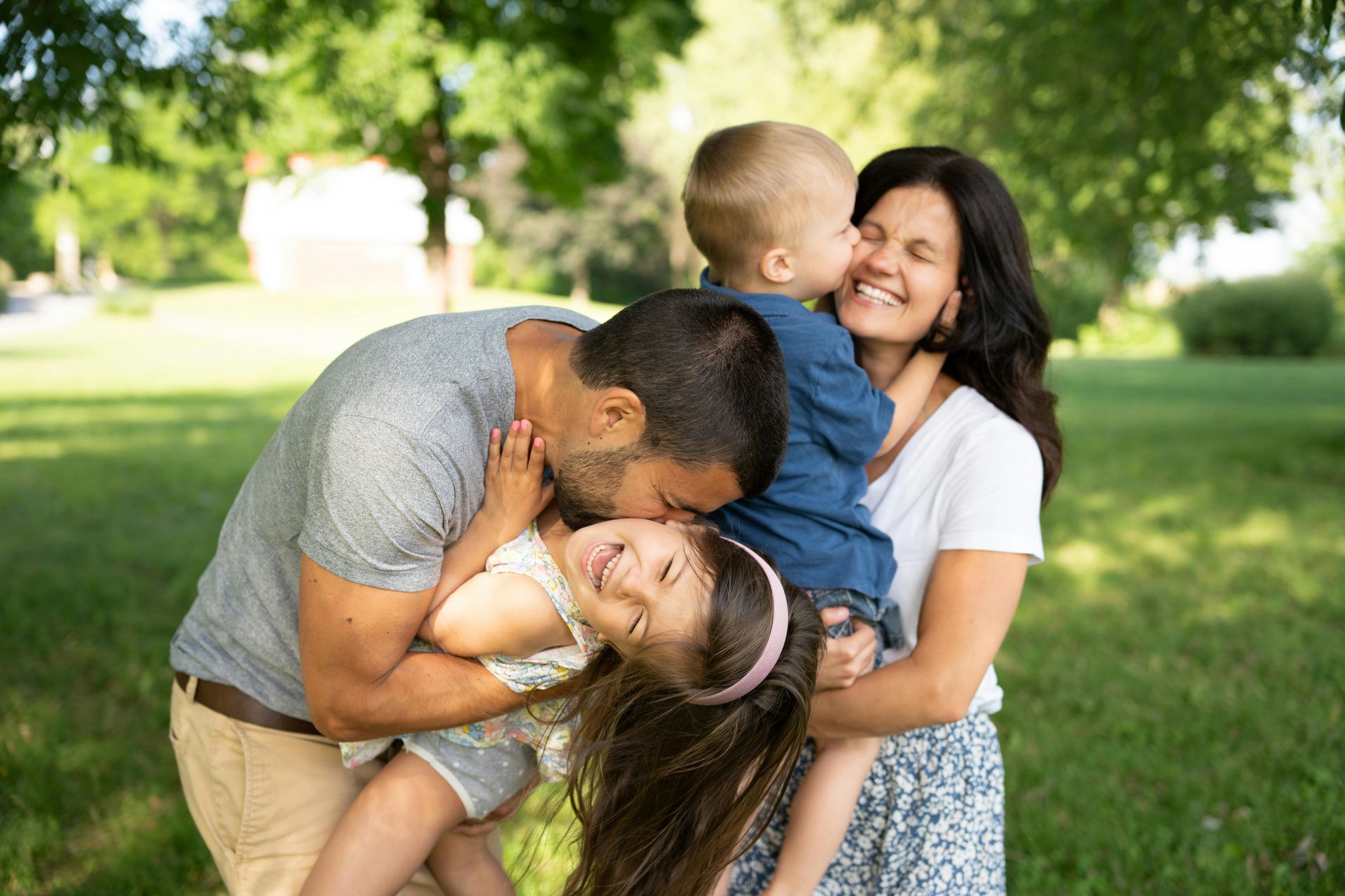 Family of four in a park, laughing. Father kissing girl, mother holding boy, all smiling.
