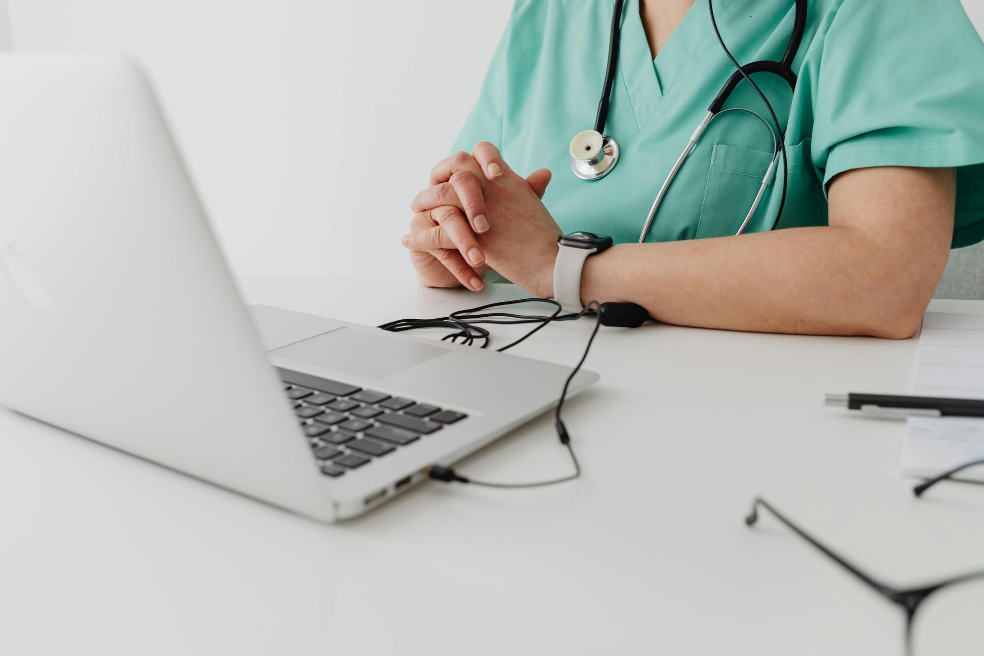Person in green scrubs sits at a desk with a laptop, stethoscope, and glasses. Hands are clasped.