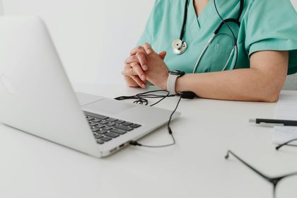 Person in green scrubs sits at a desk with a laptop, stethoscope, and glasses. Hands are clasped.