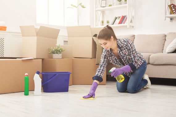 Person cleaning a living room floor with purple gloves, spray bottle, cloth, and mop near moving boxes