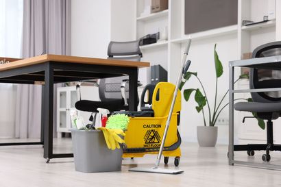 Office cleaning supplies beside a desk, with mop, bucket, caution sign, and gloves on a bright floor.