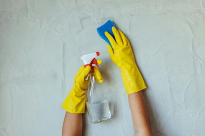 Hands in yellow gloves spraying cleaner and wiping a white wall with a blue sponge.