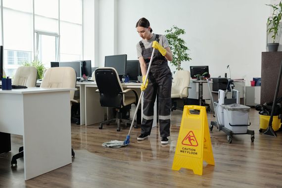 Office cleaner mopping a wood floor beside a yellow wet-floor sign in a modern workspace