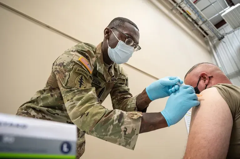 Man in military uniform administering a vaccine into the arm of another man.