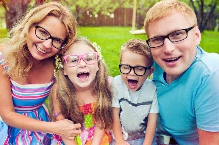 Smiling family posing outdoors in a green park, with two children wearing glasses in front.