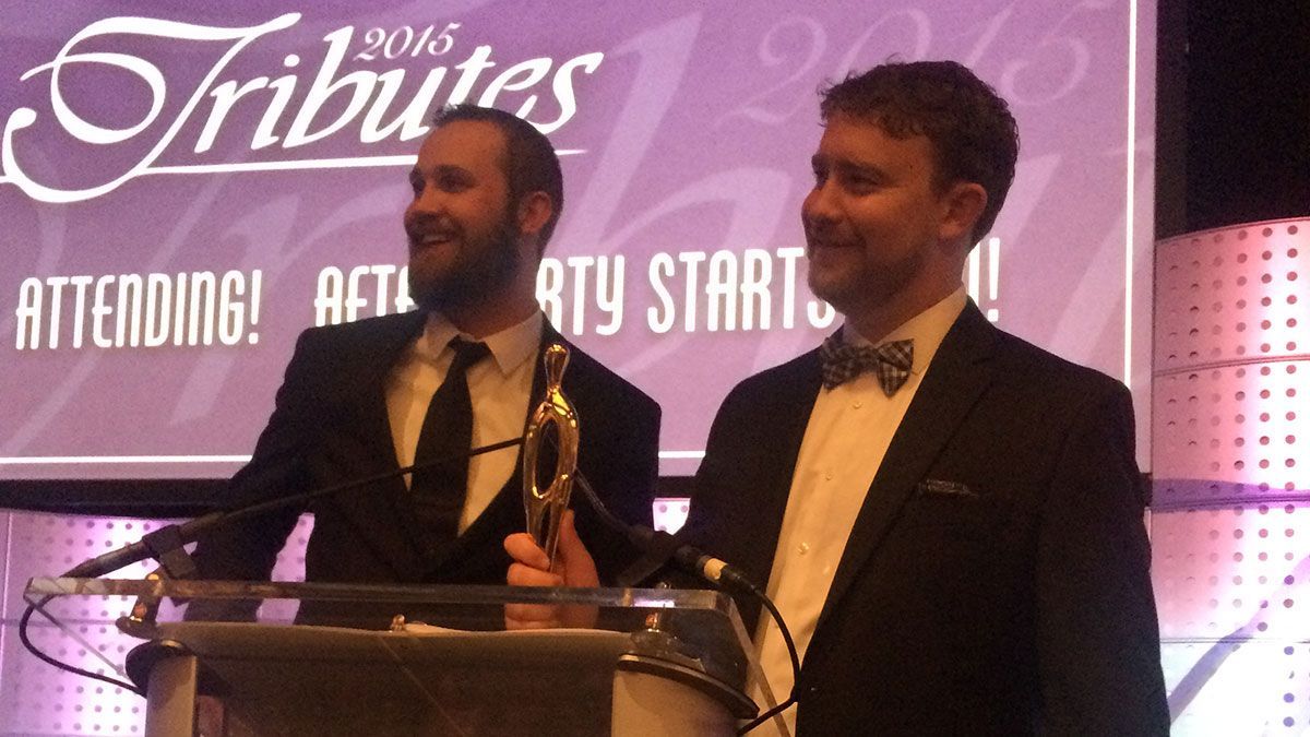 Two men standing at a podium in front of a sign that says tributes