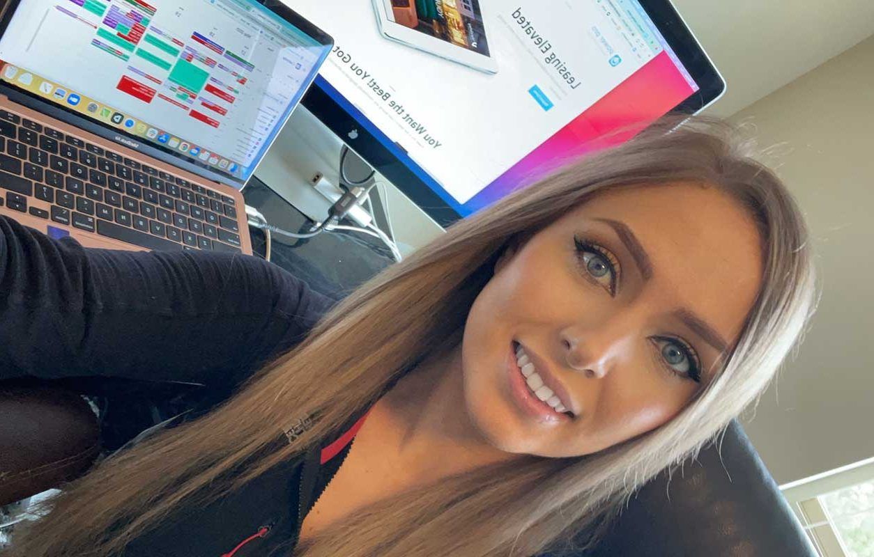 A woman is taking a selfie in front of a computer and a laptop.