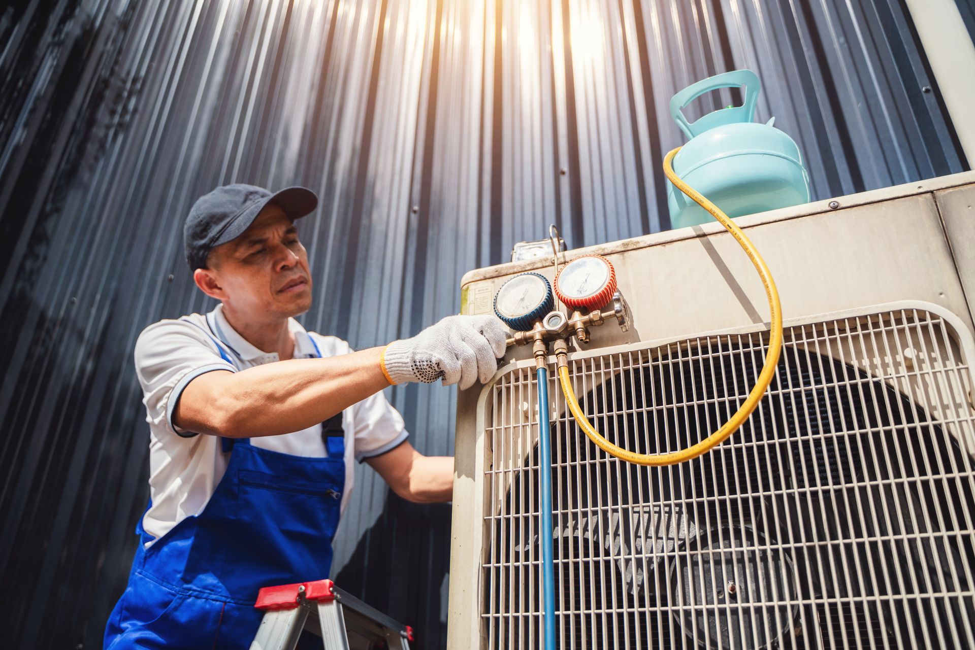 HVAC technician in blue overalls gauges an outdoor AC unit; a blue gas tank sits nearby.