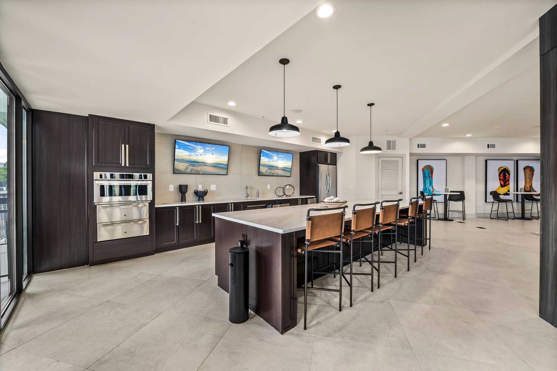 Spacious communal kitchen with dark cabinetry, large island with seating, TVs, and modern lighting.