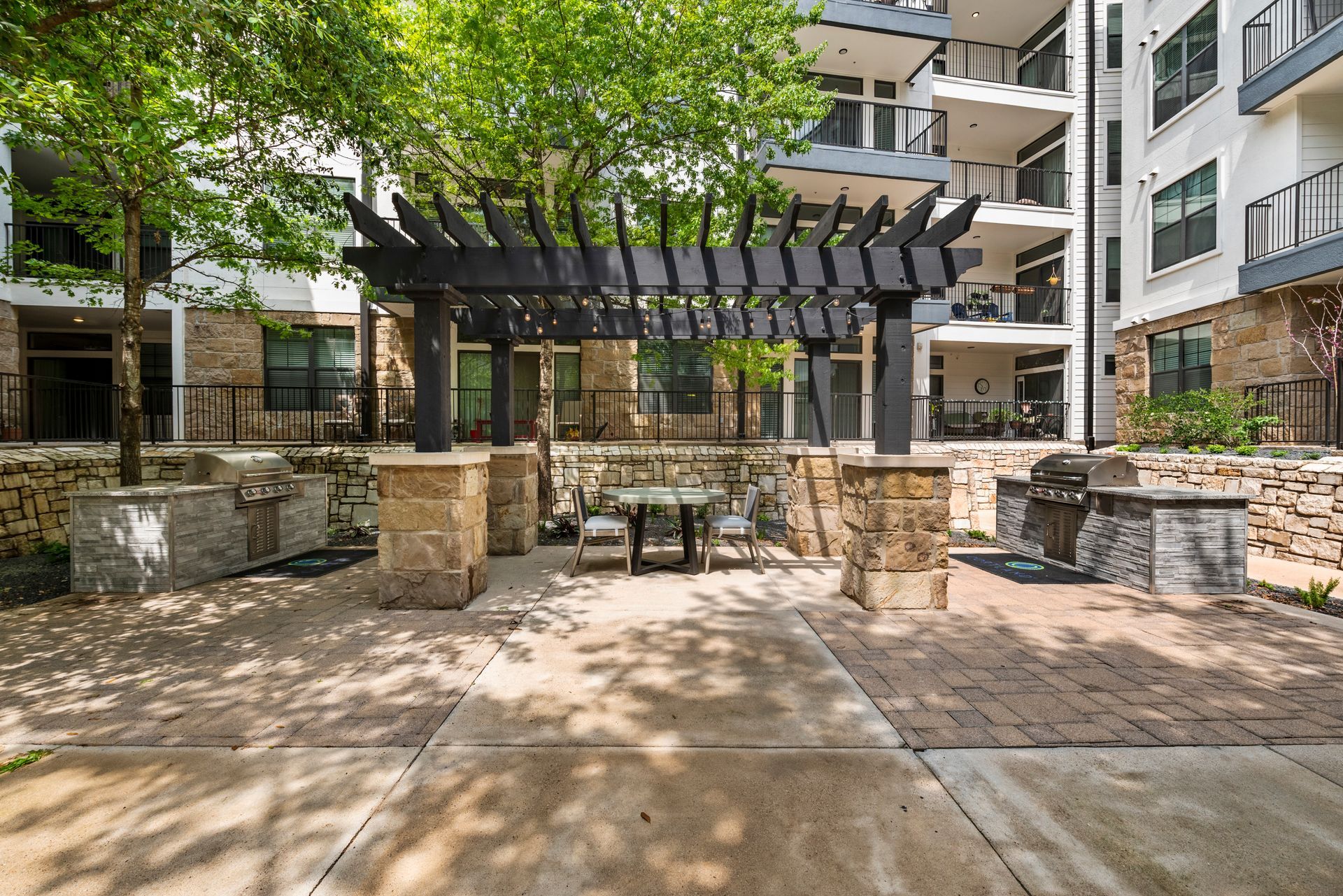 Outdoor seating area with pergola, tables, and grills between apartment buildings.