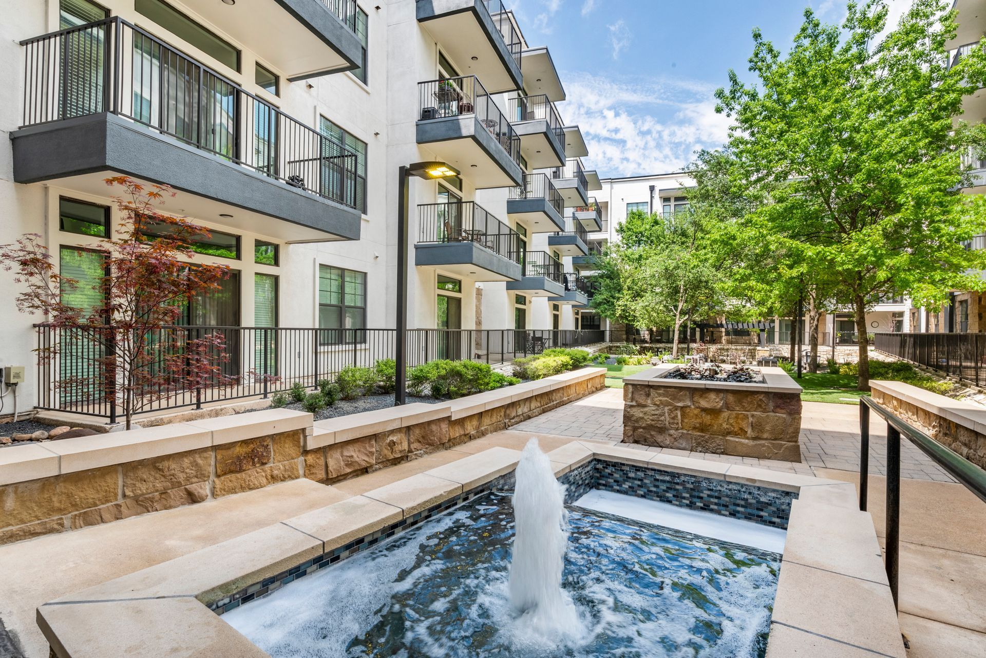 Apartment courtyard with fountain, stone accents, and trees. Multi-story building with balconies in background.