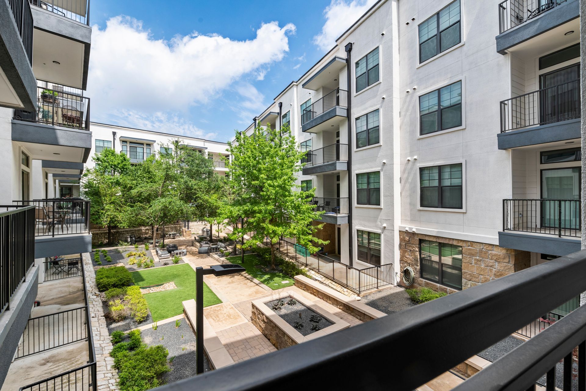 Apartment courtyard with green grass, trees, stone fire pit, and buildings with balconies on a sunny day.