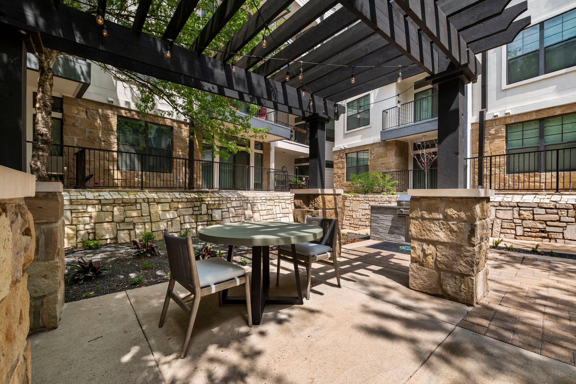 Patio with table and chairs under a black pergola. Stone walls, building in background.