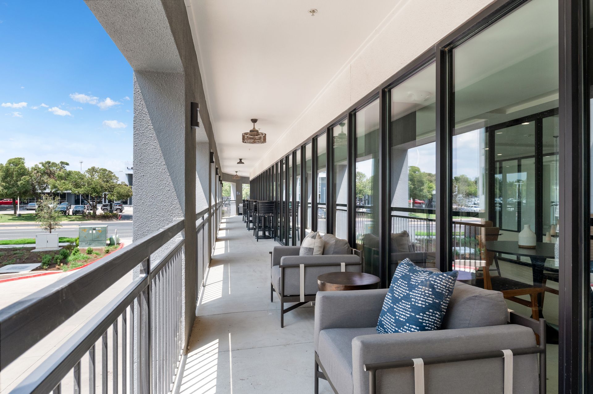 Balcony with seating, glass windows, and city view. Gray furniture, blue cushion, bright day.