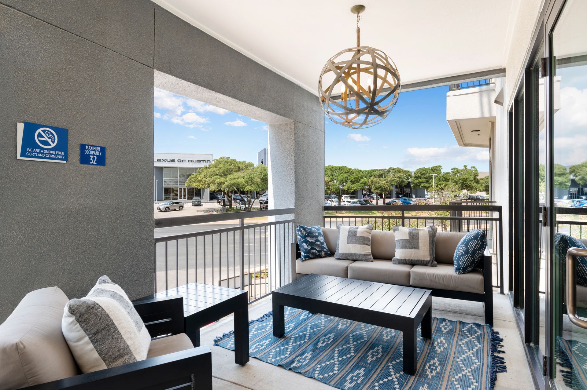 Balcony with seating: sofa, chairs, blue rug, open to cityscape with gray wall, lamp and pillows.