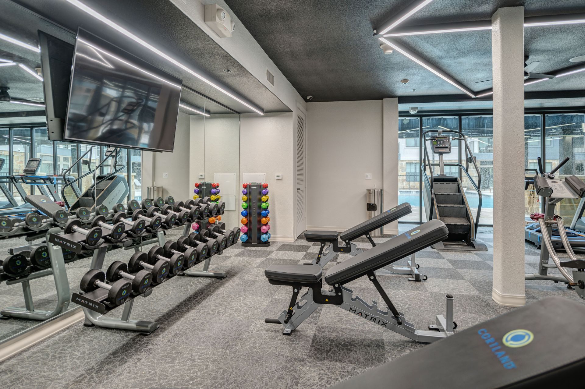 Gym interior with dumbbells, weight benches, stair climber, and exercise equipment; a mirror reflects the scene.