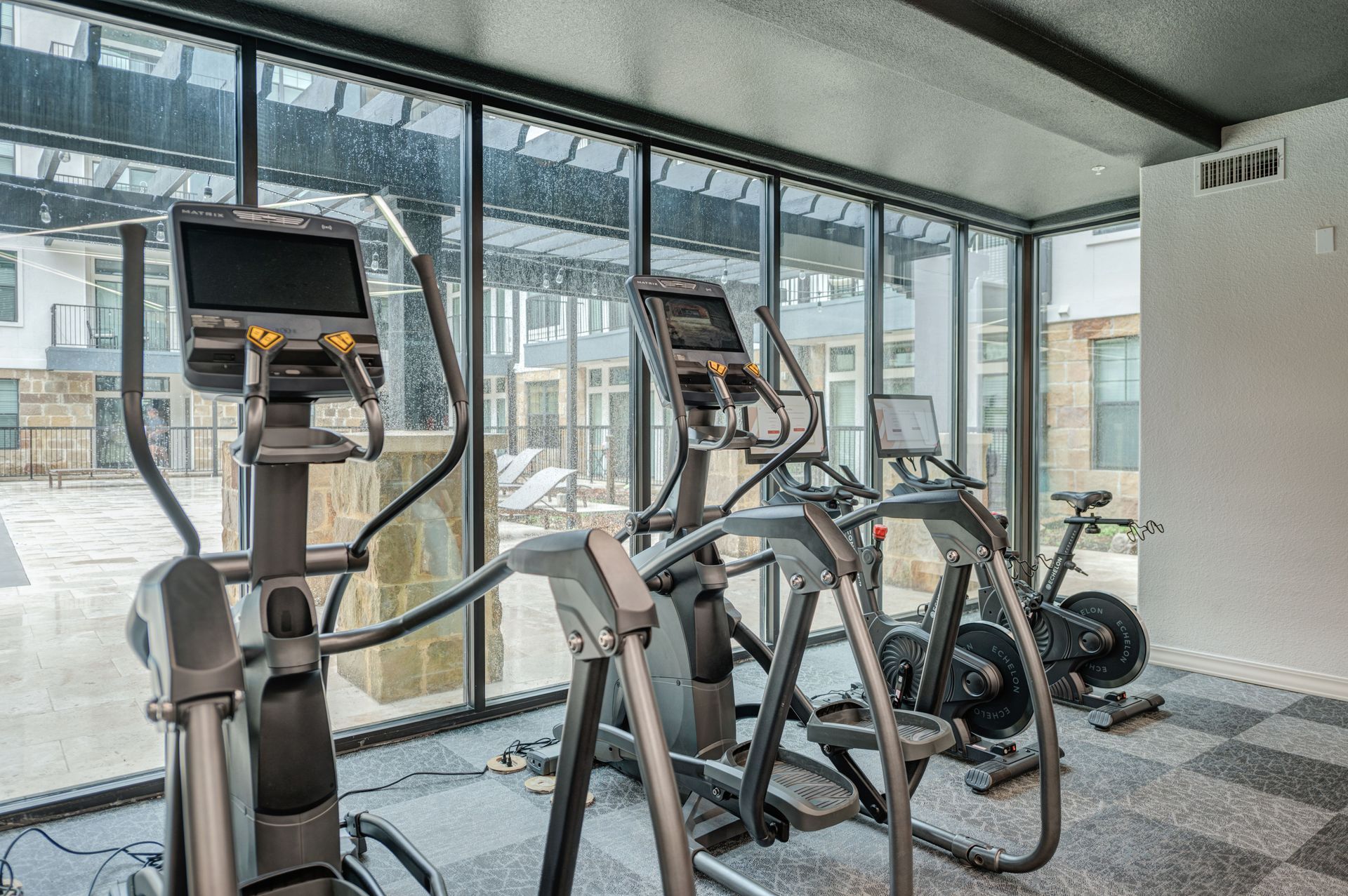 Gym with elliptical machines and stationary bike, large windows overlooking outdoor patio.