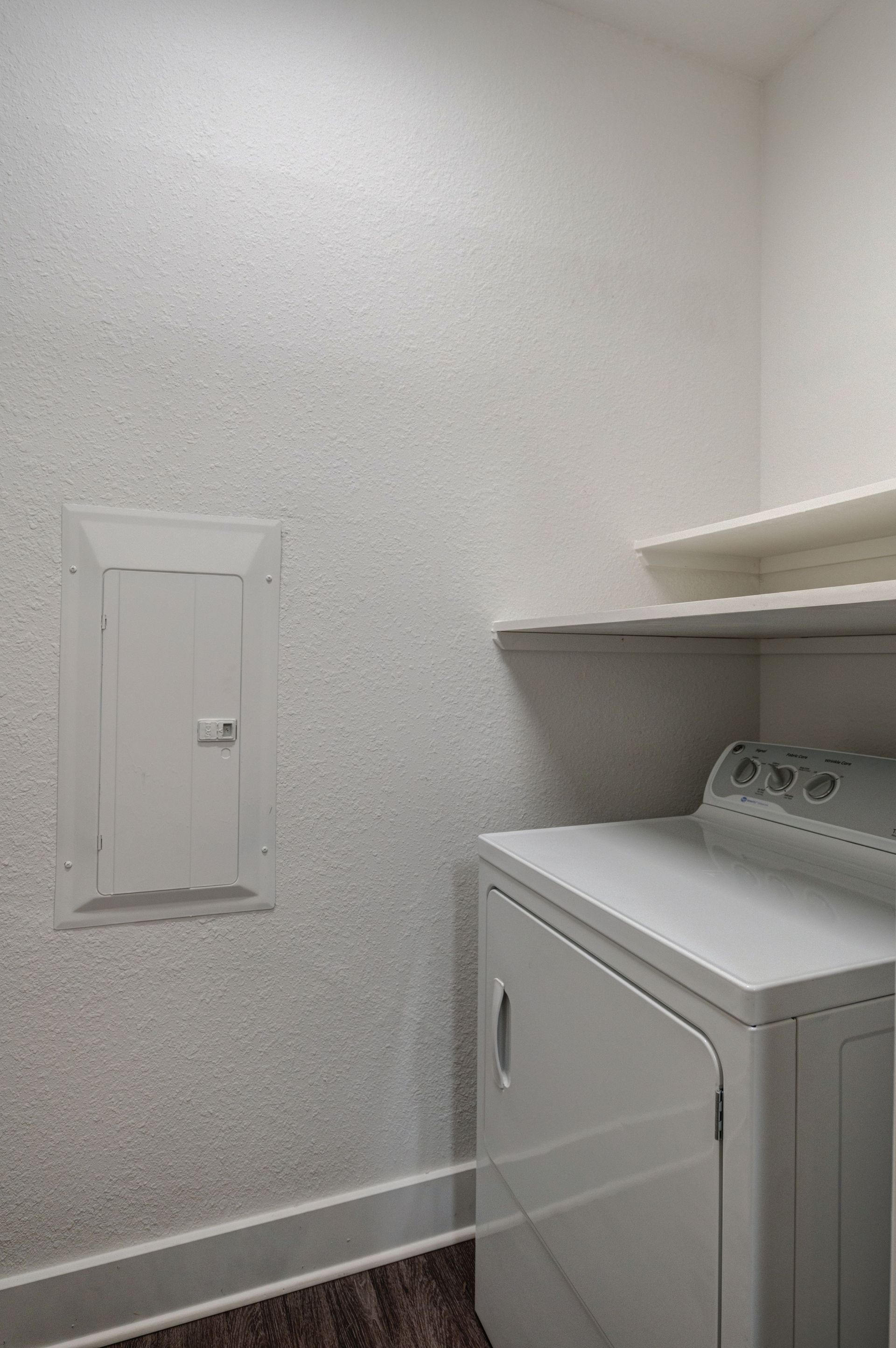 Laundry room with a white dryer, wall-mounted shelves, and an electrical panel on a textured wall.