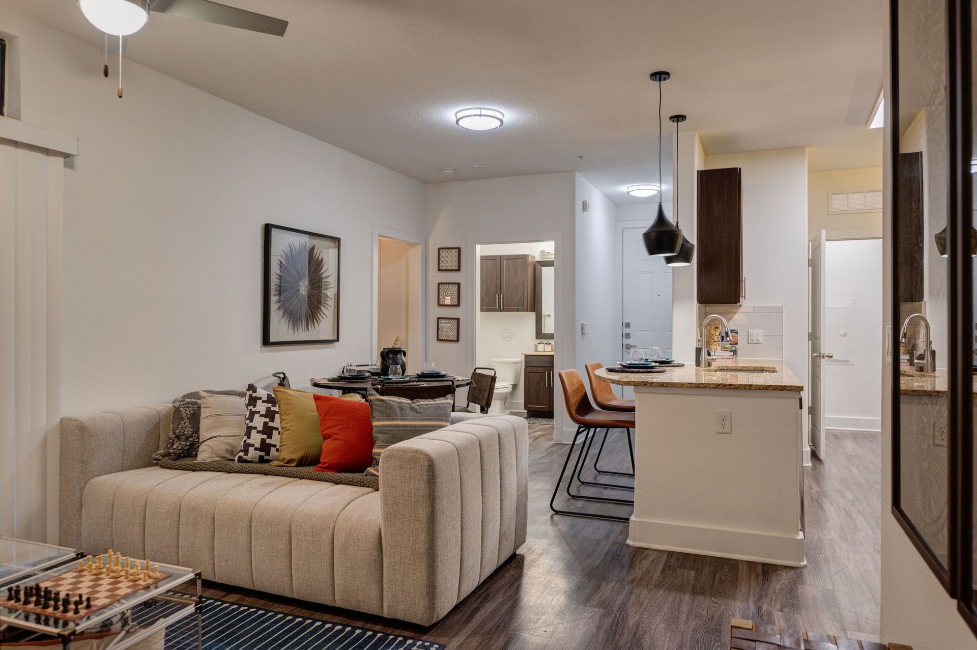 A light-filled apartment living space. Beige couch with colorful pillows, open to a kitchen with bar stools.