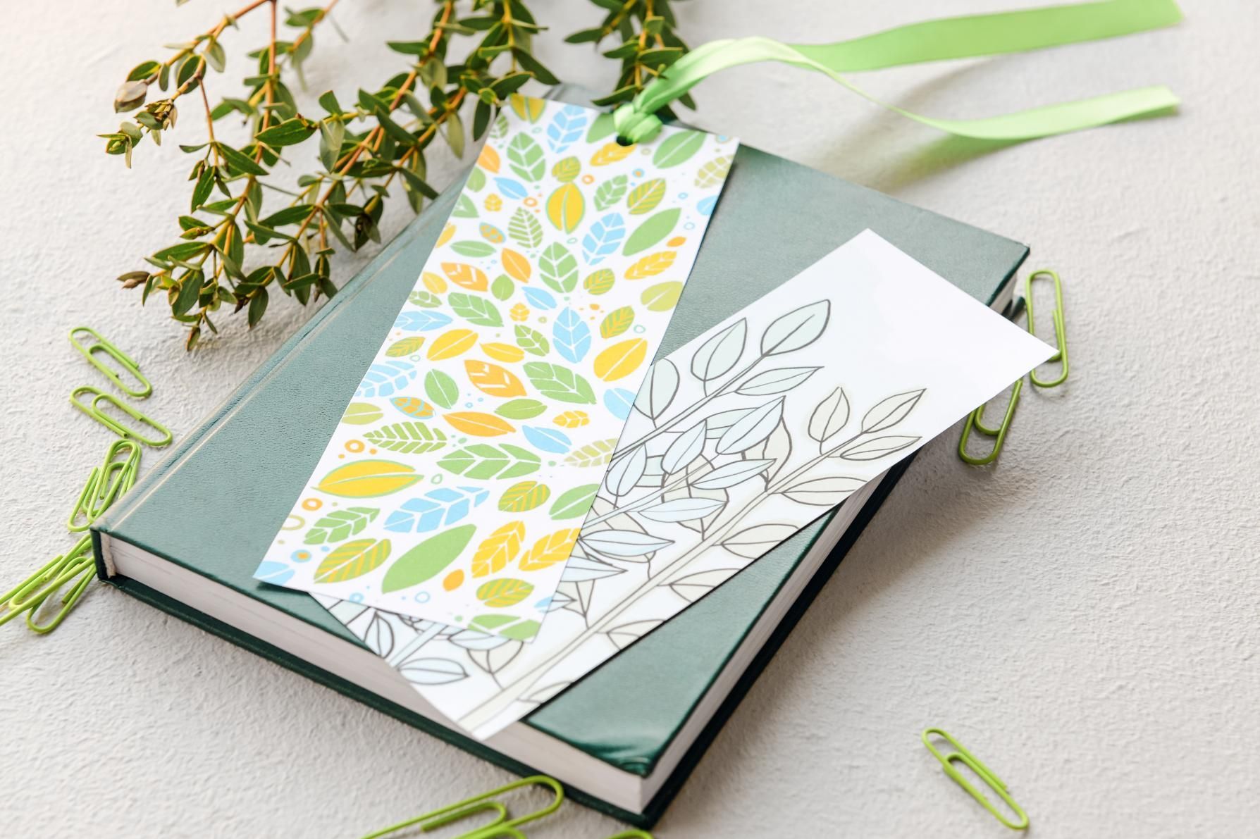 Book with two leaf-patterned bookmarks, a sprig of greenery, and green paper clips on a light surface.