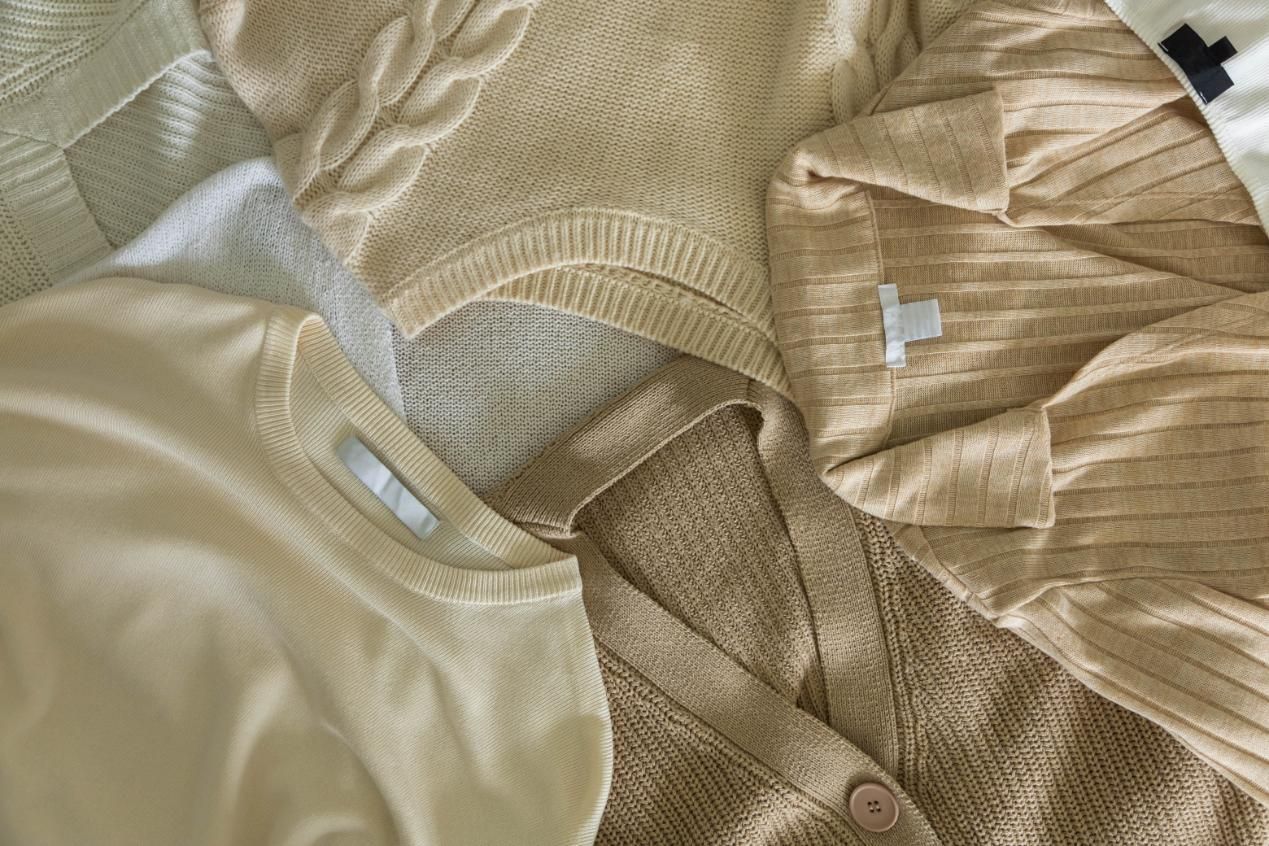 Assorted beige-toned sweaters and cardigans, various textures, overlapping on a textured surface.