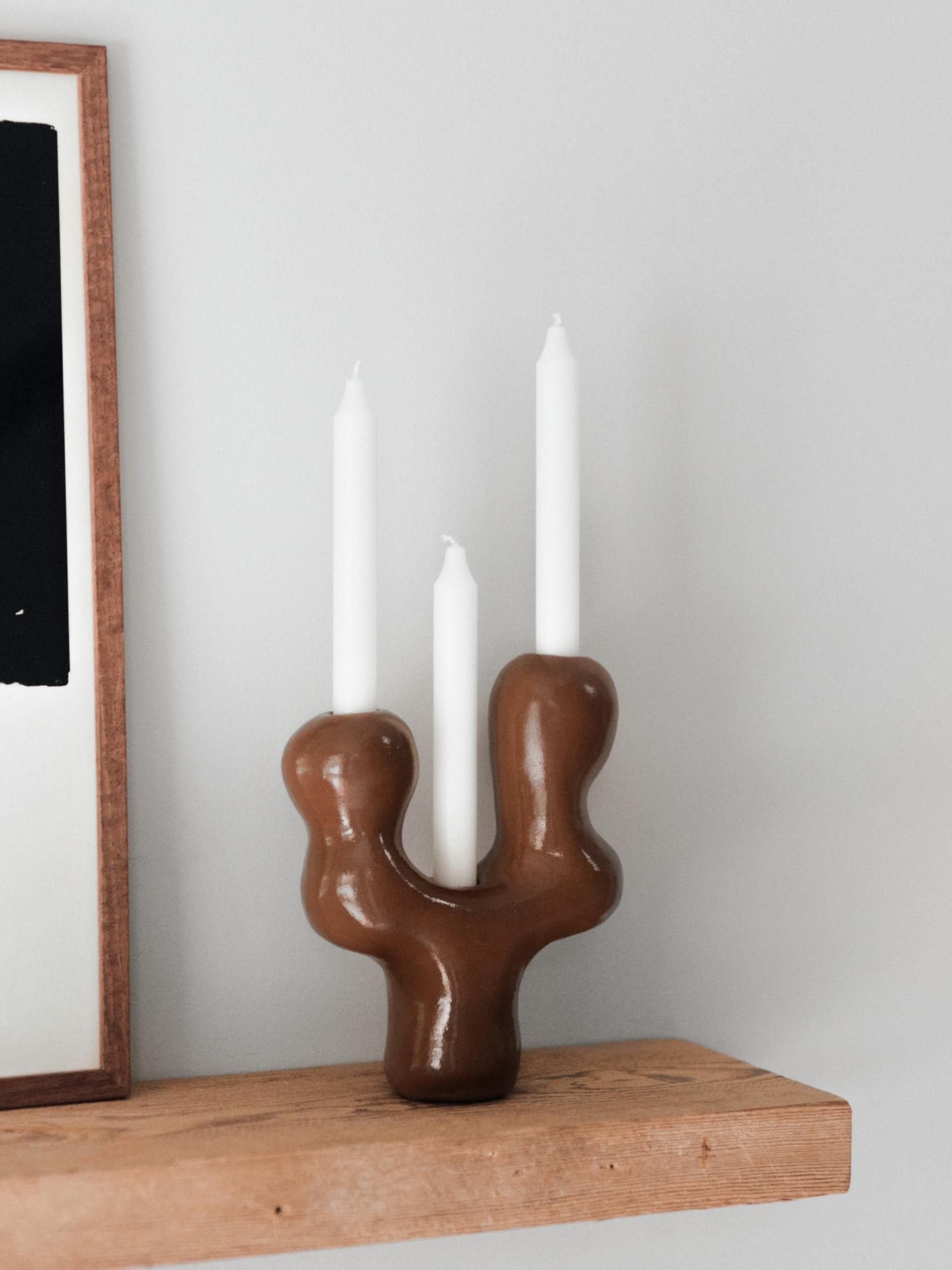 Brown candle holder with three white candles on a wooden shelf, next to a framed black and white picture.