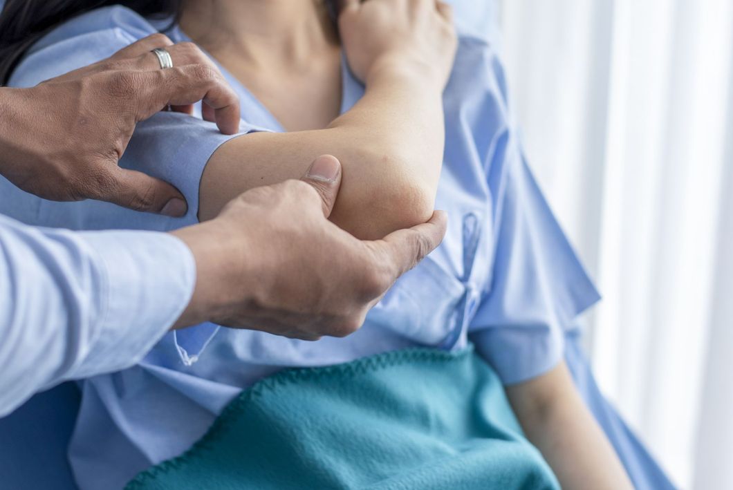 Medico che esamina il gomito di un paziente. Il paziente indossa un camice blu, all'interno di una stanza.