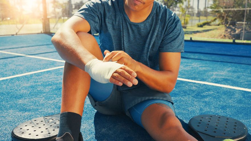 Un uomo con il polso bendato è seduto sul campo blu con le racchette da paddle; ambiente esterno soleggiato.