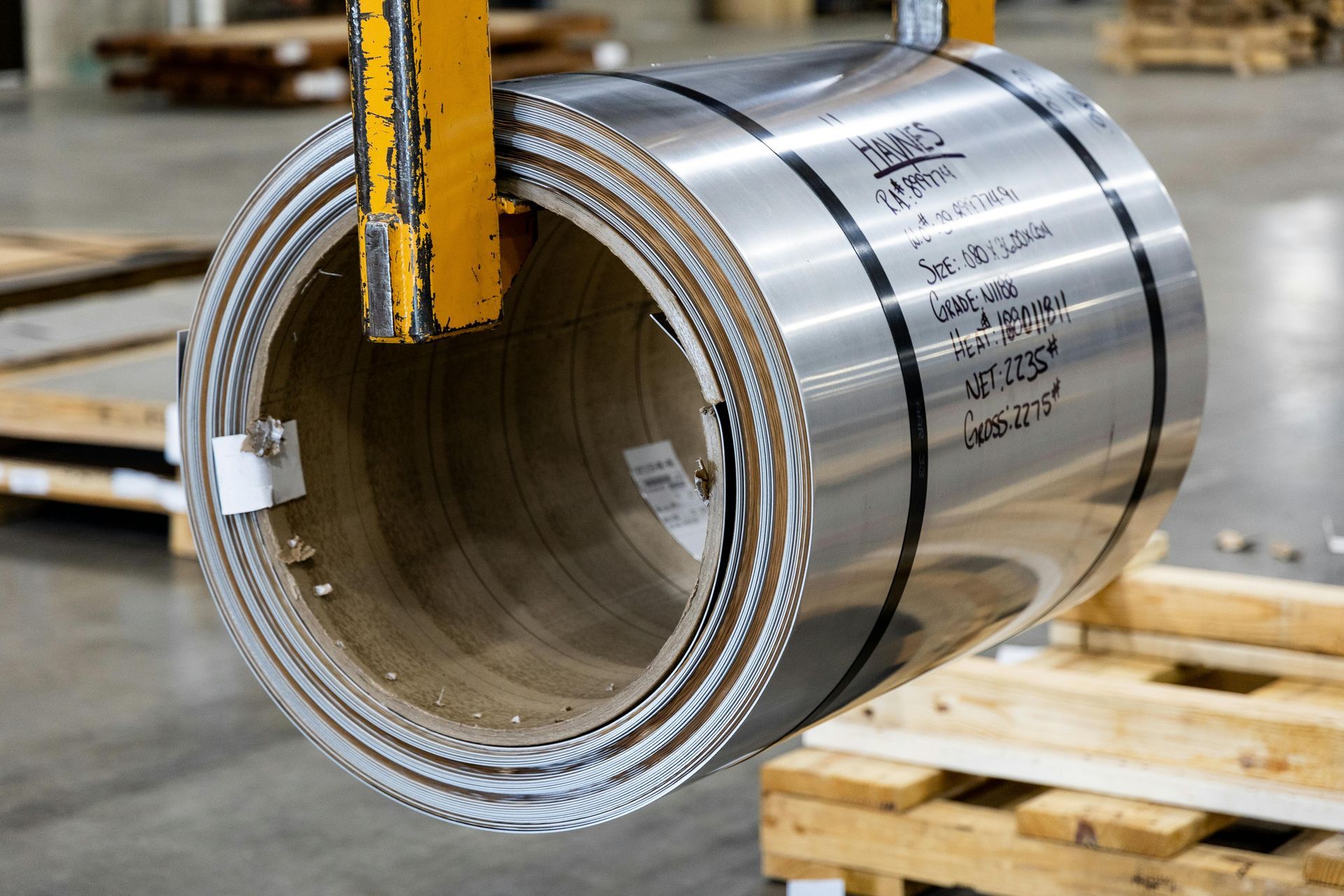 A metal coil suspended by a yellow crane, surrounded by wooden pallets in a warehouse.