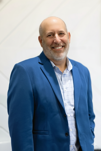 Bald man with beard smiles in a blue blazer and checkered shirt against a white backdrop.