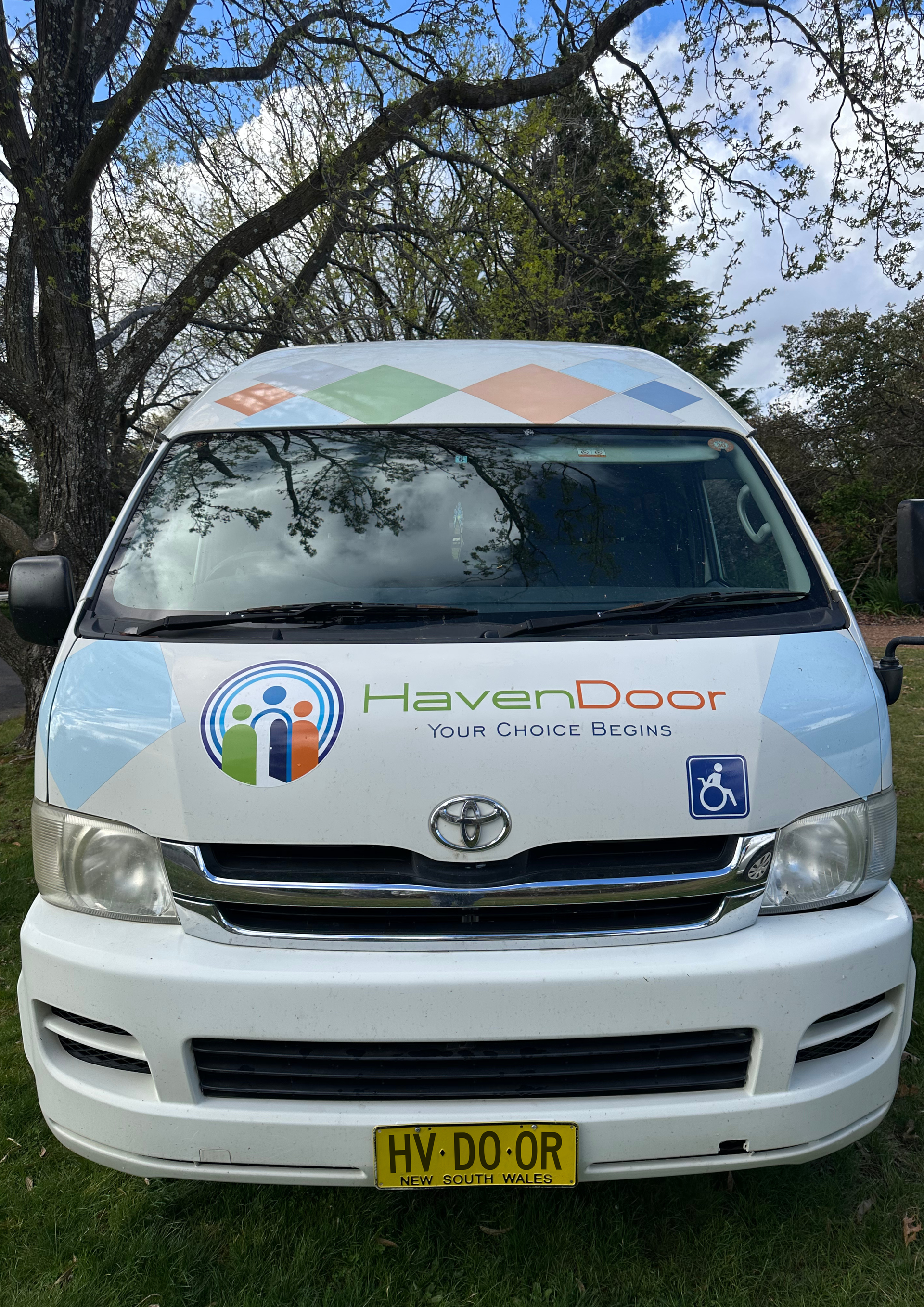 Woman helping a man in a wheelchair load a bag into a car trunk in daylight.