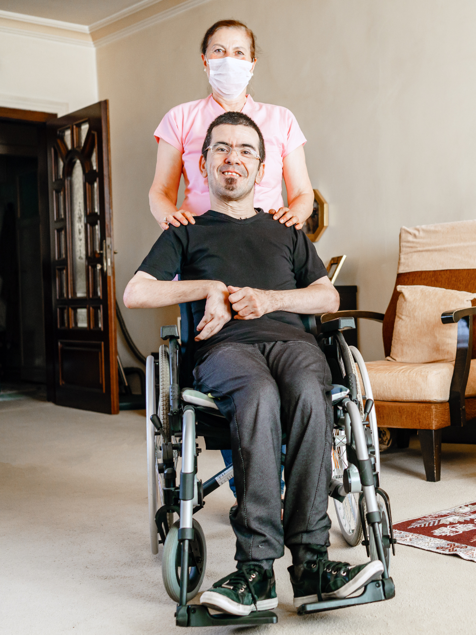 Woman helps an elderly woman from a wheelchair to a bed in a bright room.