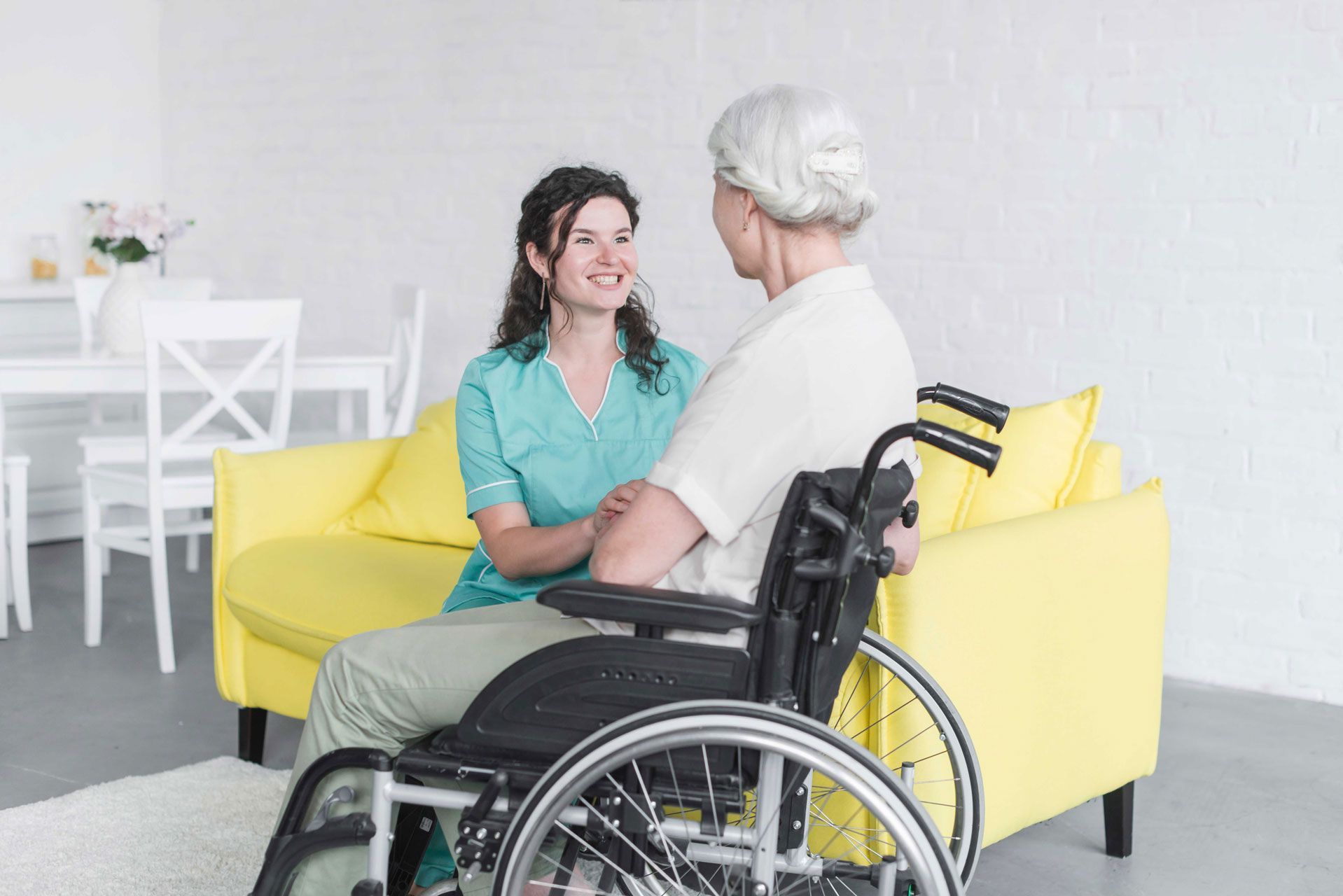 Caregiver and elderly woman in a wheelchair smiling, sitting on a yellow couch in a living room.